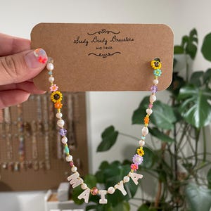 May include: A beaded necklace on a brown cardboard display card labeled Seedy Beady Bracelets AND THINGS. The necklace features white pearls, colorful flower-shaped beads, and white letter beads spelling MAIYA, interspersed with small gold-tone spacers.