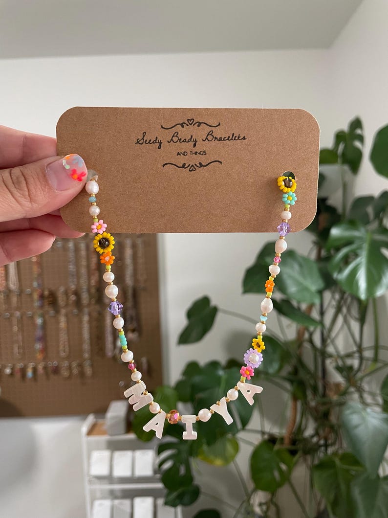 May include: A beaded necklace on a brown cardboard display card labeled Seedy Beady Bracelets AND THINGS. The necklace features white pearls, colorful flower-shaped beads, and white letter beads spelling MAIYA, interspersed with small gold-tone spacers.