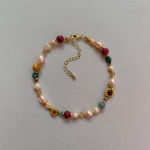 May include: A beaded necklace featuring irregular white freshwater pearls, round red and yellow beads, and small flower-shaped beads in teal and yellow. The necklace includes gold-toned metal spacers and a gold-toned lobster clasp with an extender chain.