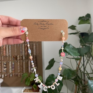 May include: A beaded necklace featuring a mix of pastel-colored round beads, small flower-shaped beads, and white letter beads spelling out NICOLE. The necklace is attached to a brown rectangular display card with the text Seedy Beady Bracelets.