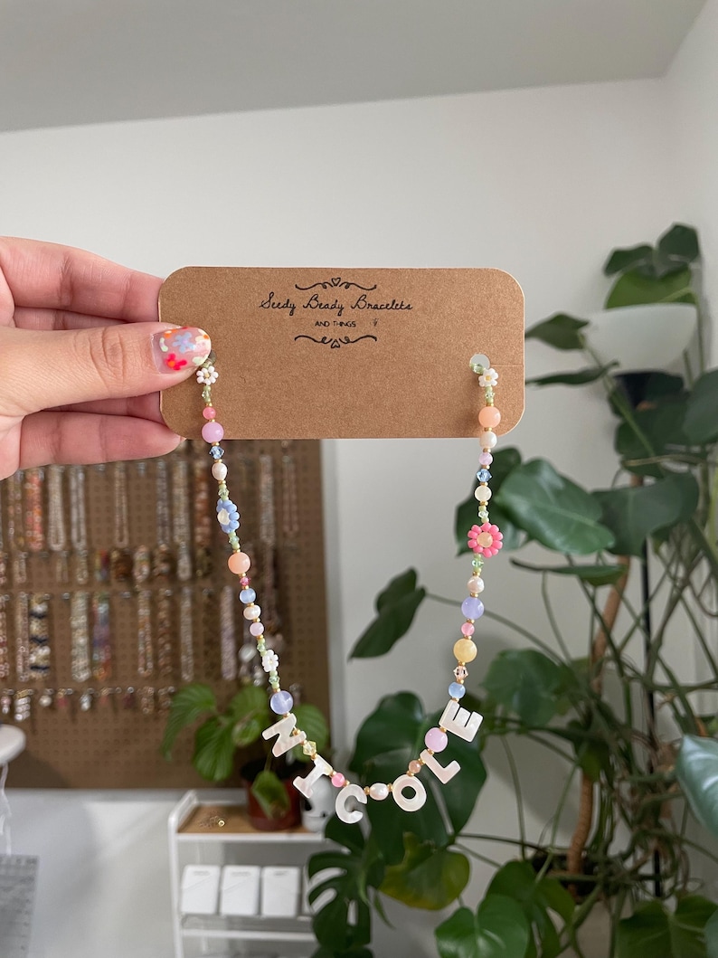 May include: A beaded necklace featuring a mix of pastel-colored round beads, small flower-shaped beads, and white letter beads spelling out NICOLE. The necklace is attached to a brown rectangular display card with the text Seedy Beady Bracelets.