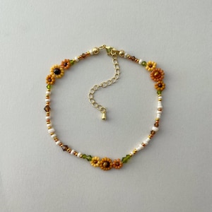 May include: A beaded bracelet featuring a floral design with yellow and orange sunflowers. The bracelet includes white, gold, and brown beads, with green accents. The clasp is gold-colored.