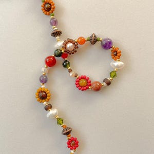 May include: A colorful beaded necklace featuring a variety of beads in shades of red, orange, yellow, purple, and green. The necklace includes flower-shaped beads, pearls, and gold-colored accents. The design is intricate and eye-catching, perfect for adding a pop of color to any outfit.