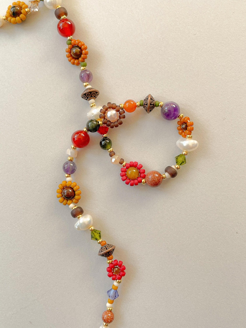May include: A colorful beaded necklace featuring a variety of beads in shades of red, orange, yellow, purple, and green. The necklace includes flower-shaped beads, pearls, and gold-colored accents. The design is intricate and eye-catching, perfect for adding a pop of color to any outfit.