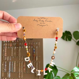 May include: A beaded necklace featuring a mix of small colorful glass beads, brown flower-shaped beads, and white letter beads spelling the name DELILAH. The necklace is attached to a rectangular brown cardboard display card labeled Seedy Beady Bracelets.