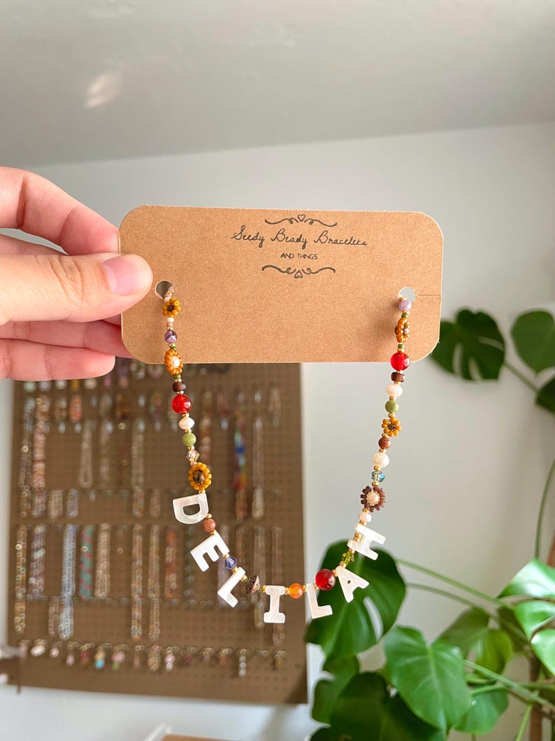 May include: A beaded necklace featuring a mix of small colorful glass beads, brown flower-shaped beads, and white letter beads spelling the name DELILAH. The necklace is attached to a rectangular brown cardboard display card labeled Seedy Beady Bracelets.