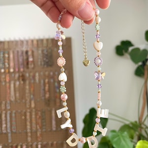 May include: A beaded necklace featuring white mother-of-pearl letter beads spelling LYDIE, interspersed with pastel-colored flower, butterfly, and heart-shaped beads, small round pearls, and translucent purple beads, finished with a gold-tone metal chain.