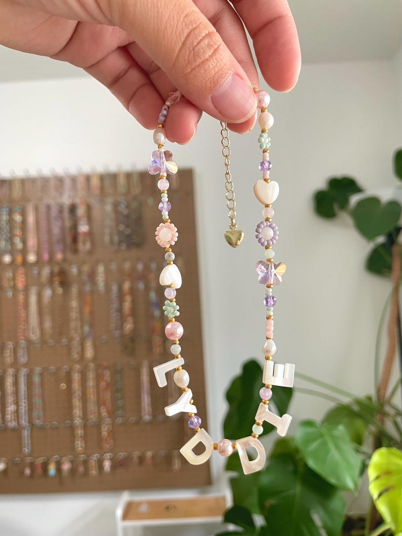 May include: A beaded necklace featuring white mother-of-pearl letter beads spelling LYDIE, interspersed with pastel-colored flower, butterfly, and heart-shaped beads, small round pearls, and translucent purple beads, finished with a gold-tone metal chain.