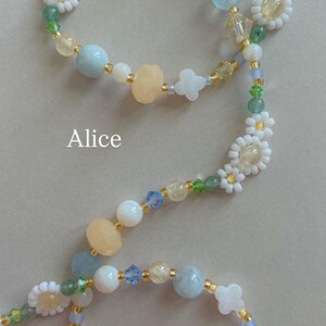 May include: A beaded accessory featuring the name Alice, crafted with light blue, pale yellow, and white beads, interspersed with small green accents and white flower-shaped beads. The design includes faceted crystals and smooth round beads on a thin cord.