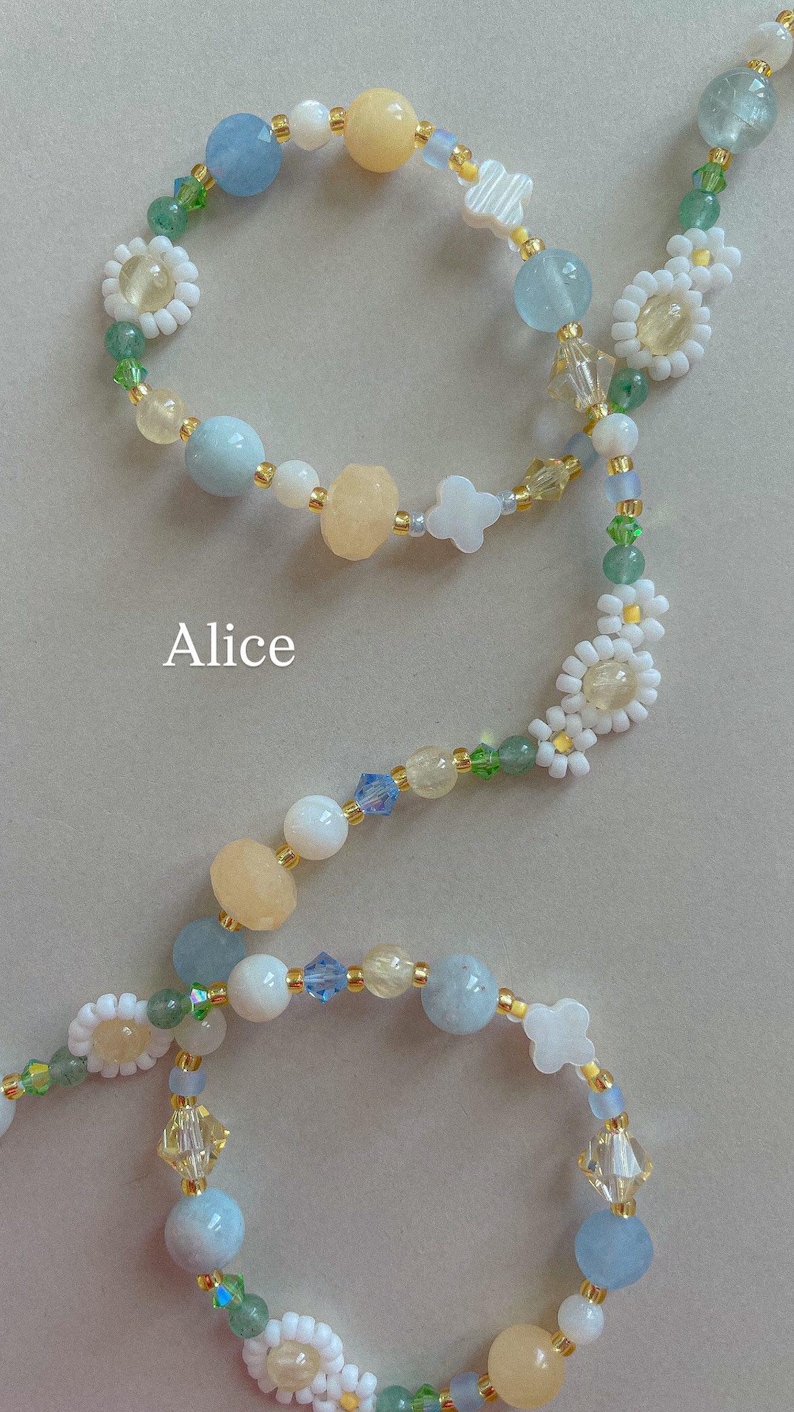 May include: A beaded accessory featuring the name Alice, crafted with light blue, pale yellow, and white beads, interspersed with small green accents and white flower-shaped beads. The design includes faceted crystals and smooth round beads on a thin cord.