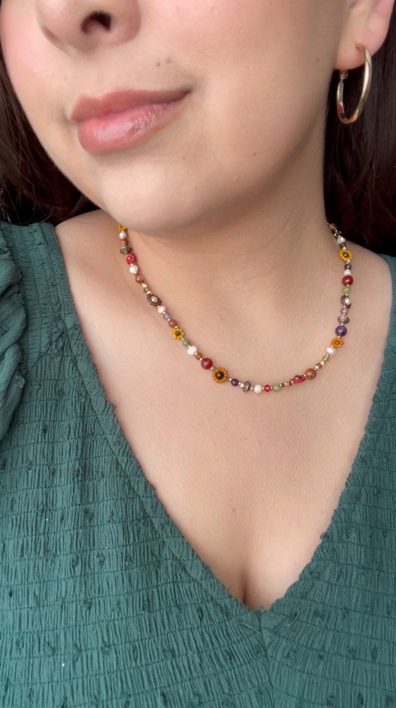 May include: A colorful beaded necklace with a variety of beads in red, yellow, white, and green. The necklace features small flower-shaped beads and gold-colored metal accents. The person is wearing a green top and gold hoop earrings.