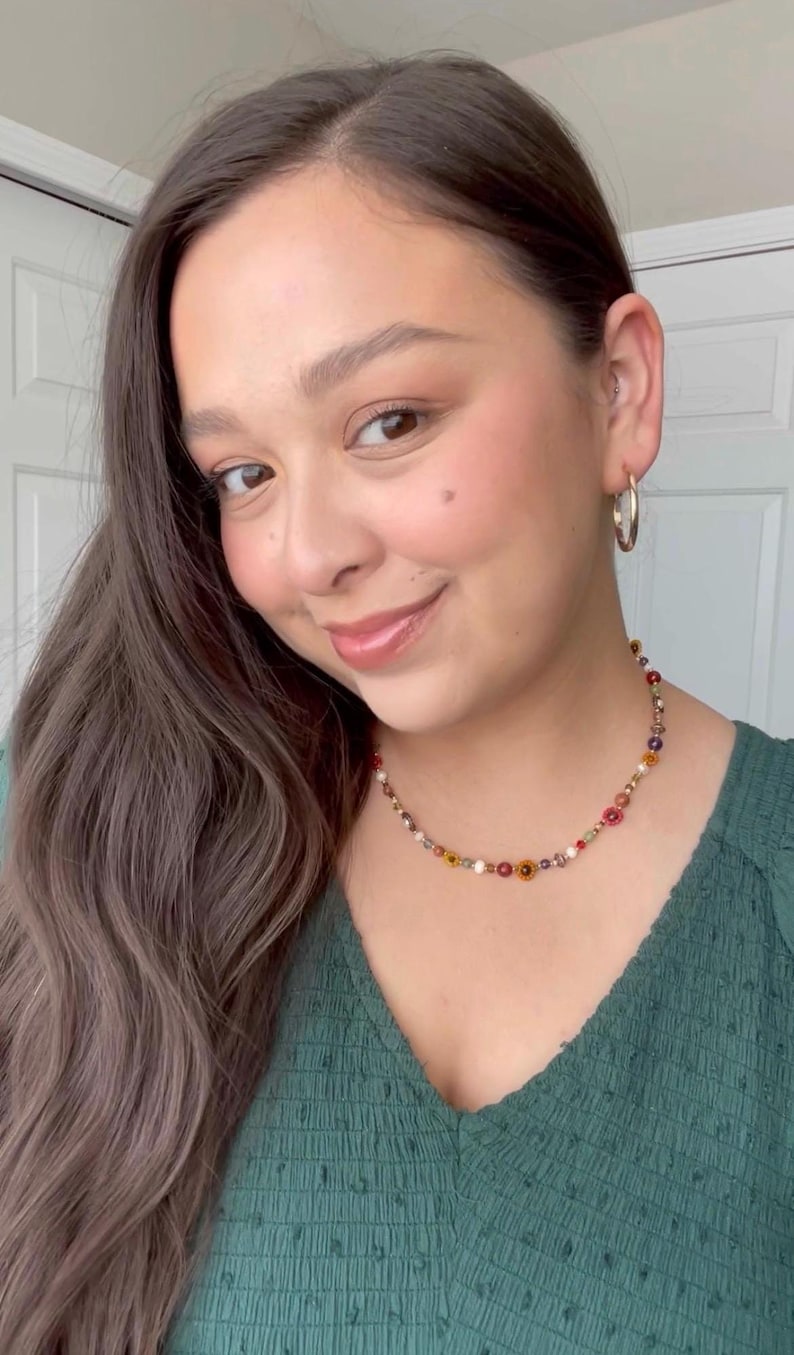 May include: A colorful beaded necklace with small flower and bead accents. The necklace features a variety of colors, including red, yellow, and white. The woman is wearing a green top and silver hoop earrings.