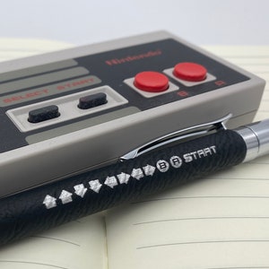 May include: A black pen with a silver tip and a textured grip. The pen has the words "B A START" printed on it. The pen is lying on top of a gray Nintendo controller.