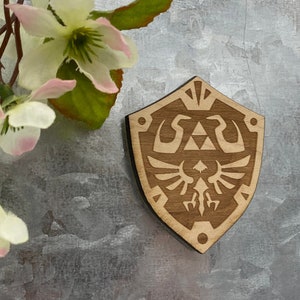 May include: A wooden shield with a brown stain, featuring a stylized design of the Triforce and a winged creature. The shield is a popular symbol from the Legend of Zelda video game series.