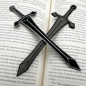 May include: Two black sword bookmarks crossed over a book. The swords have a silver-grey outline and a black blade.
