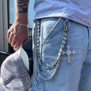 May include: A silver chain belt with a decorative cross pendant hangs from the waistband of light blue denim shorts. A matching silver bracelet is on the wrist. A white and gray trucker hat is held in the hand.