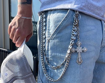 May include: A silver chain belt with a decorative cross pendant hangs from the waistband of light blue denim shorts. A matching silver bracelet is on the wrist. A white and gray trucker hat is held in the hand.