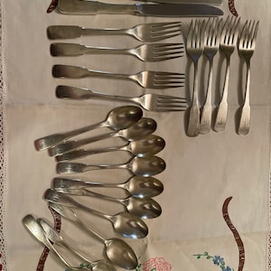 May include: A set of silverware, including spoons, forks, and knives, laid out on a white tablecloth with floral embroidery. The silverware is made of polished silver and has a traditional design.