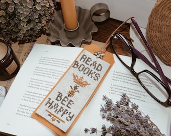 May include: A close-up of a book with a bookmark that says "Read Books Bee Happy". The bookmark is made of fabric and has a bee design. The book is open to a page with text in a foreign language. There are also other items in the image, including a candle, a plant, and a pair of glasses.