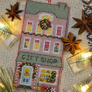 May include: A pink and green cross-stitch ornament depicting a gift shop with a wreath above the door and a garland of beads around the windows. The ornament is on a burlap background with lights and pine branches.