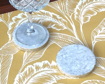 May include: Two round, white, marble coasters with a gold glitter edge. The coasters are on a yellow background with a white leaf pattern.