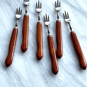 Può includere: Sette forchette da portata con rebbi in acciaio inossidabile e manici in legno marrone scuro. Le forchette sono disposte su una superficie grigio chiaro, mostrando il loro design elegante. Un set di utensili da tavola.