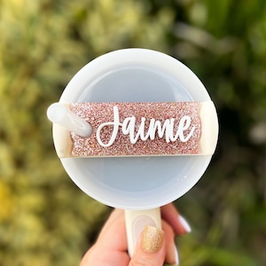 May include: A white and light blue fan with a handle, featuring a glittery rose gold band with the name "Jaime" in white script. The fan is held against a blurred green background.