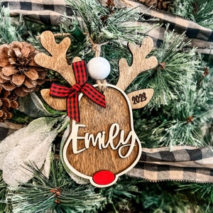 May include: Wooden reindeer Christmas ornament with a red and black plaid bow, a white pom-pom, and the name "Emily" and the year "2023" engraved on it.