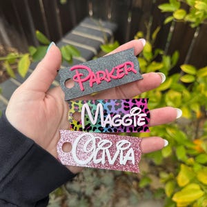 May include: Three personalized keychains with names: "Parker" in pink, "Maggie" in white, and "Olivia" in white. The keychains have a tag shape with a hole for attachment. The "Maggie" keychain has a colorful leopard print background.