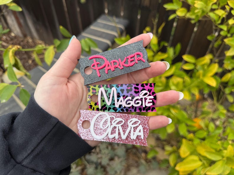 May include: Three personalized keychains with names: "Parker" in pink, "Maggie" in white, and "Olivia" in white. The keychains have a tag shape with a hole for attachment. The "Maggie" keychain has a colorful leopard print background.