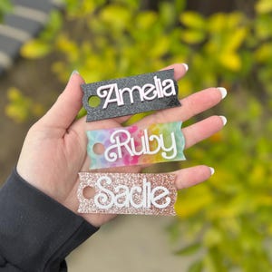 May include: Three personalized name tags with the names "Amelia", "Ruby", and "Sadie". The "Amelia" tag is black with white lettering, "Ruby" is on a tie-dye background, and "Sadie" is glittery rose gold. Each tag has a small hole.