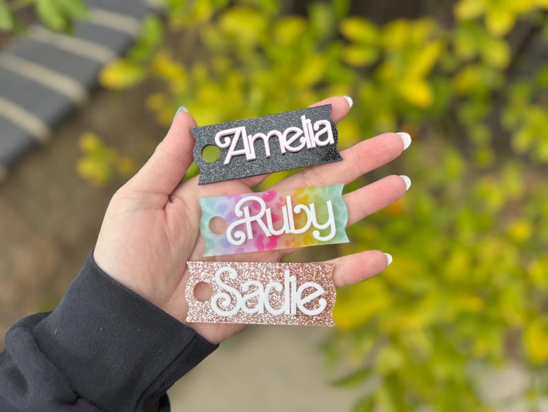 May include: Three personalized name tags with the names "Amelia", "Ruby", and "Sadie". The "Amelia" tag is black with white lettering, "Ruby" is on a tie-dye background, and "Sadie" is glittery rose gold. Each tag has a small hole.