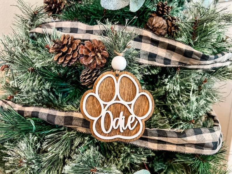 May include: A wooden Christmas ornament in the shape of a dog paw print, with the name "Odie" in a cursive font. The ornament is hanging on a Christmas tree decorated with pine needles, pine cones, and a black and white plaid ribbon.