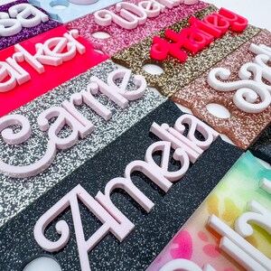 May include: A collection of personalized name tags in various colors and glitter finishes. Each tag features a different name in a contrasting color, with names like "Amelia", "Carrie", and "Haley" visible. The tags are likely for events or gifts.
