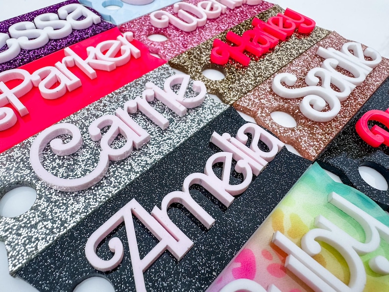 May include: A collection of personalized name tags in various colors and glitter finishes. Each tag features a different name in a contrasting color, with names like "Amelia", "Carrie", and "Haley" visible. The tags are likely for events or gifts.