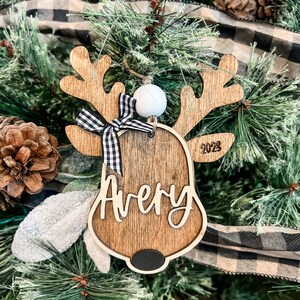 May include: Wooden reindeer ornament with a black and white checkered bow, a white bead, and the name "Avery" and the year "2023" engraved on it.