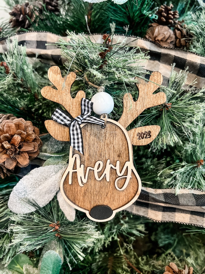 May include: Wooden reindeer ornament with a black and white checkered bow, a white bead, and the name "Avery" and the year "2023" engraved on it.