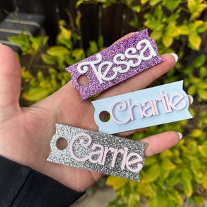 May include: Three glittery name tags are displayed. The tags are rectangular with a decorative edge and a hole. The names "Tessa", "Charlie", and "Carrie" are written on the tags in white and pink lettering. The tags are purple, light blue, and silver.