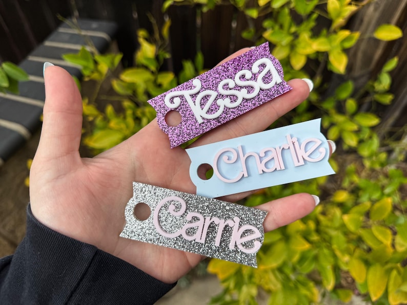 May include: Three glittery name tags are displayed. The tags are rectangular with a decorative edge and a hole. The names "Tessa", "Charlie", and "Carrie" are written on the tags in white and pink lettering. The tags are purple, light blue, and silver.