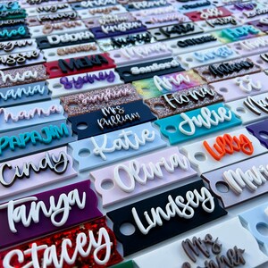 Op de afbeelding: Een verzameling kleurrijke gepersonaliseerde naamstickers met verschillende lettertypen en stijlen. De stickers zijn gemaakt van acryl en hebben een gat om op te hangen. Sommige van de namen zijn "Olivia", "Katie", "Julie", "Tanya", "Stacey", "Lindsey", "Papajim", "Mama", "Shana", "Cindy" en "Mrs. Pulliam".