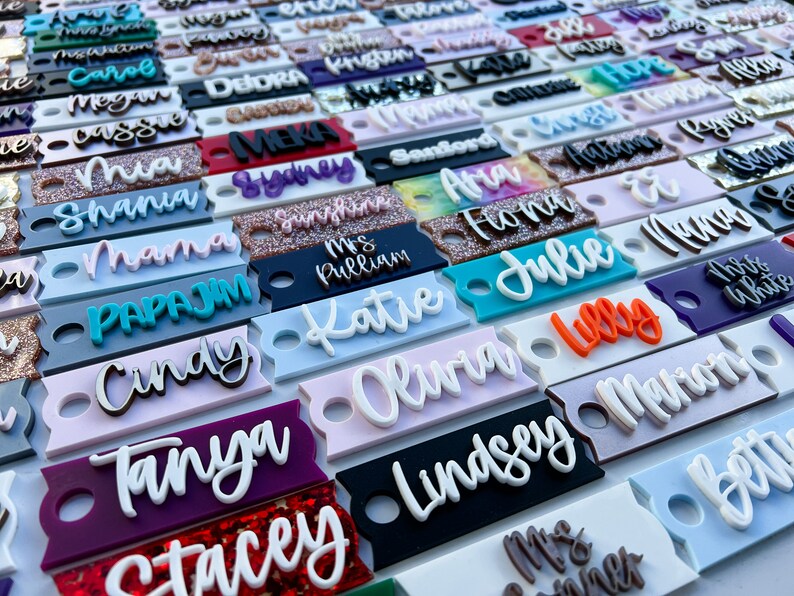 Op de afbeelding: Een verzameling kleurrijke gepersonaliseerde naamstickers met verschillende lettertypen en stijlen. De stickers zijn gemaakt van acryl en hebben een gat om op te hangen. Sommige van de namen zijn "Olivia", "Katie", "Julie", "Tanya", "Stacey", "Lindsey", "Papajim", "Mama", "Shana", "Cindy" en "Mrs. Pulliam".