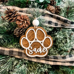 May include: A wooden paw print ornament with the name "Sadie" cut out in white. The ornament hangs on a Christmas tree with pine needles, pine cones, and a black and white plaid ribbon.
