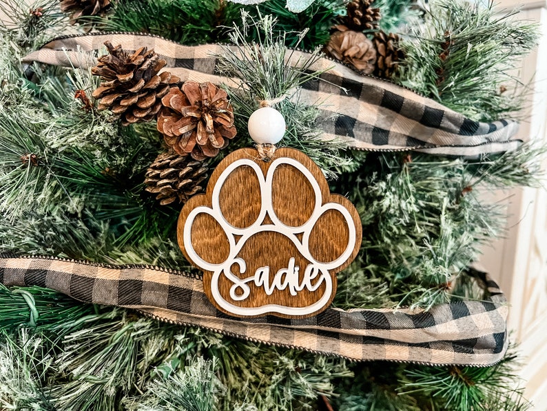 May include: A wooden paw print ornament with the name "Sadie" cut out in white. The ornament hangs on a Christmas tree with pine needles, pine cones, and a black and white plaid ribbon.