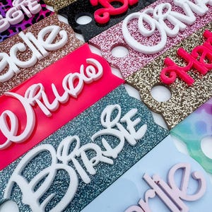 May include: Close-up of several name tags with glittered backgrounds in various colors, including pink, black, and gold. The names are in white and pink raised lettering. The tags are for a bridal party.