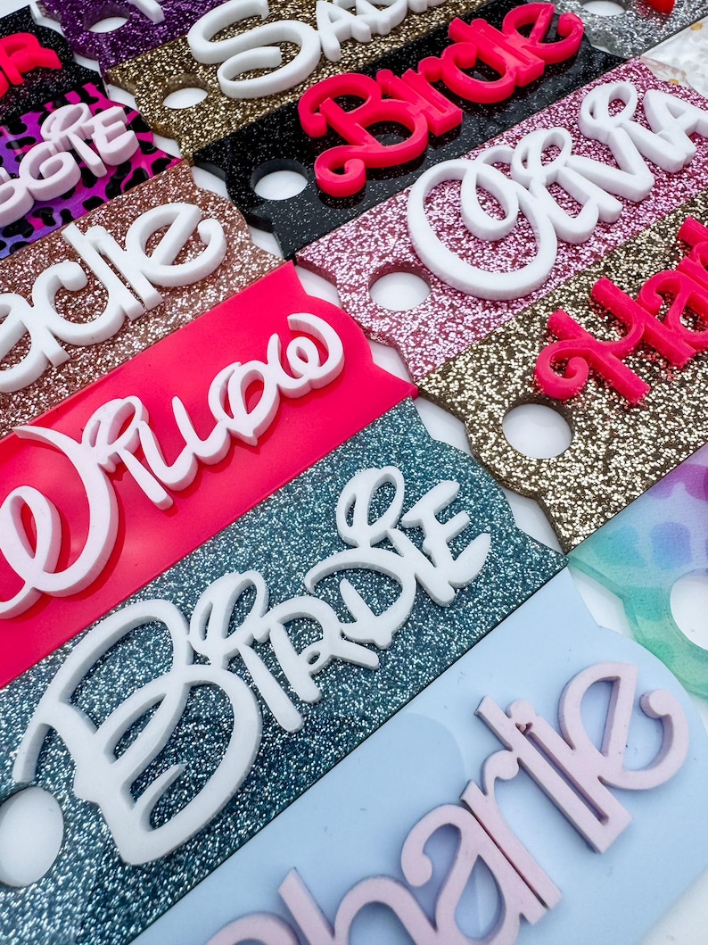 May include: Close-up of several name tags with glittered backgrounds in various colors, including pink, black, and gold. The names are in white and pink raised lettering. The tags are for a bridal party.