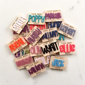 Op de afbeelding: Een verzameling houten naamlabels met diverse namen in kleurrijke, verhoogde letters. Elk label heeft de tekst "LOOK WHAT MADE" erop gedrukt. De namen zijn onder meer "Olivia", "Poppy", "Parker", "Abigail", "Mary", "Sullivan", "Wyatt", "Elle", "Jack" en anderen.