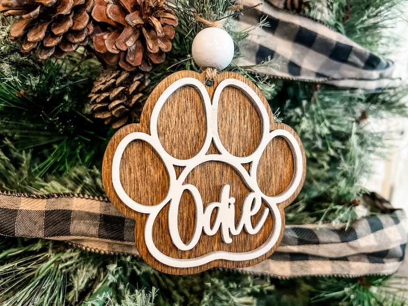 May include: A wooden Christmas ornament in the shape of a dog paw print. The ornament is brown with a white outline and the name "Odie" in a cursive font. It hangs from a white bead and is decorated with pine needles and a buffalo check ribbon.