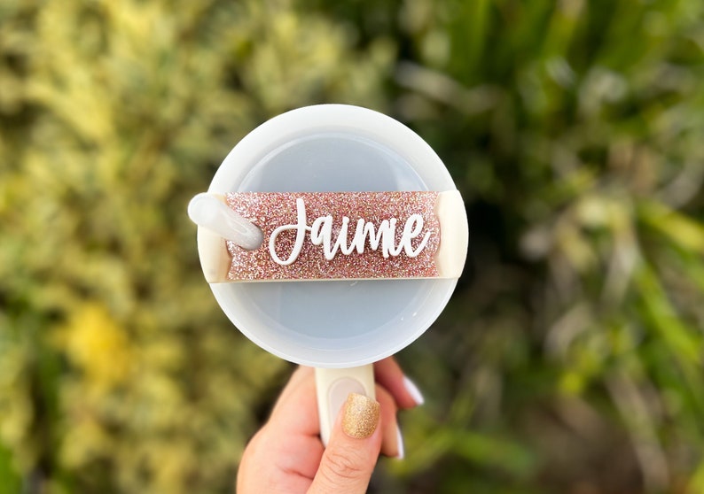 May include: A white and light blue fan with a handle, featuring a glittery rose gold band with the name "Jaime" in white script. The fan is held against a blurred green background.