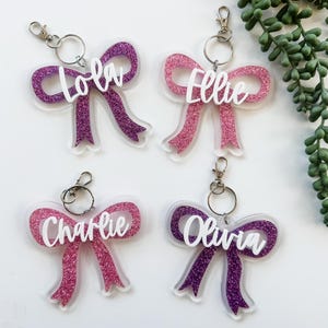 May include: Four glitter bow keychains in pink and purple with names: Lola, Ellie, Charlie, and Olivia. Each keychain has a silver clasp and ring. The bows are on a clear background.