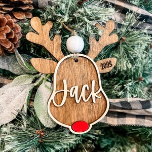 May include: Wooden reindeer ornament with a red nose, white bead, and the name "Jack" written on it. The ornament also includes the year "2023" on the side.
